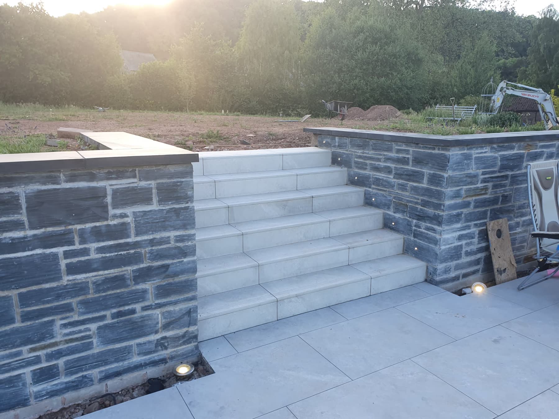 Illuminated stone-clad steps and retaining wall beside a paved patio at dusk