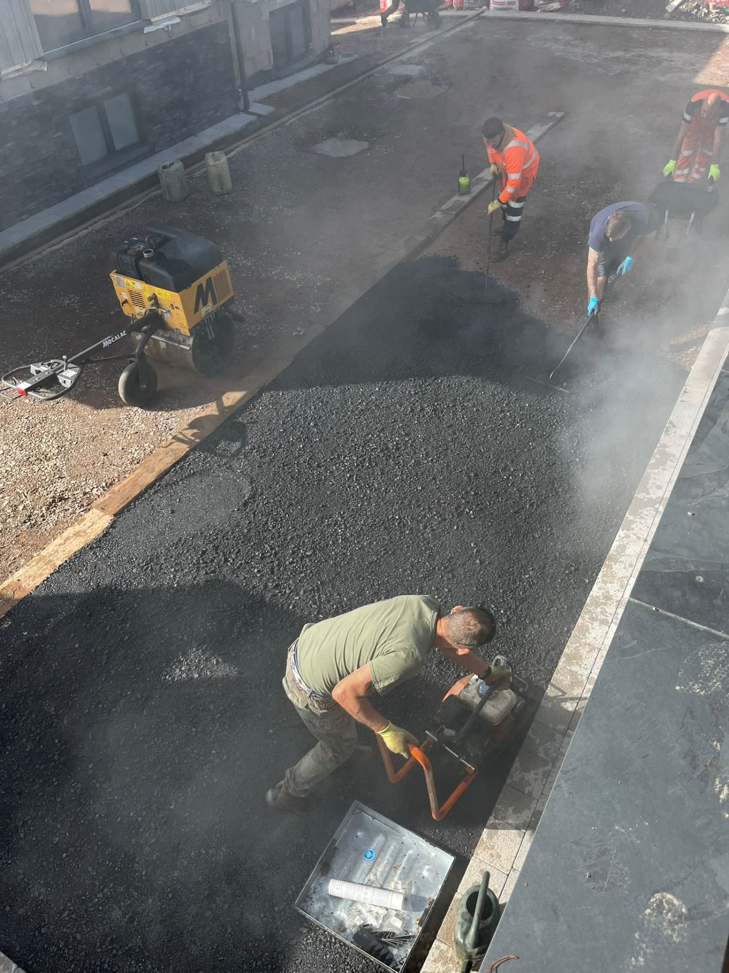 Workers compacting and finishing fresh tarmac beside a newly paved courtyard