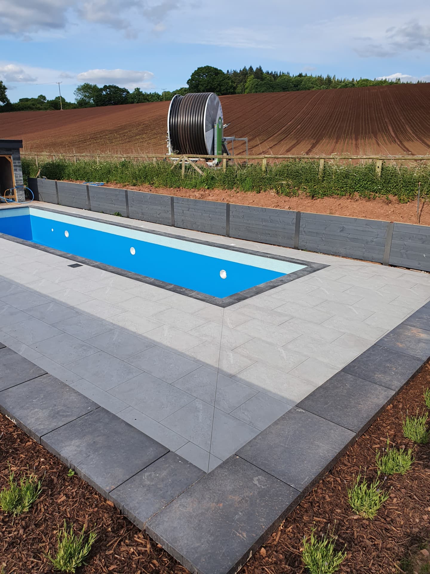Rectangular outdoor swimming pool with grey paving overlooking open farmland