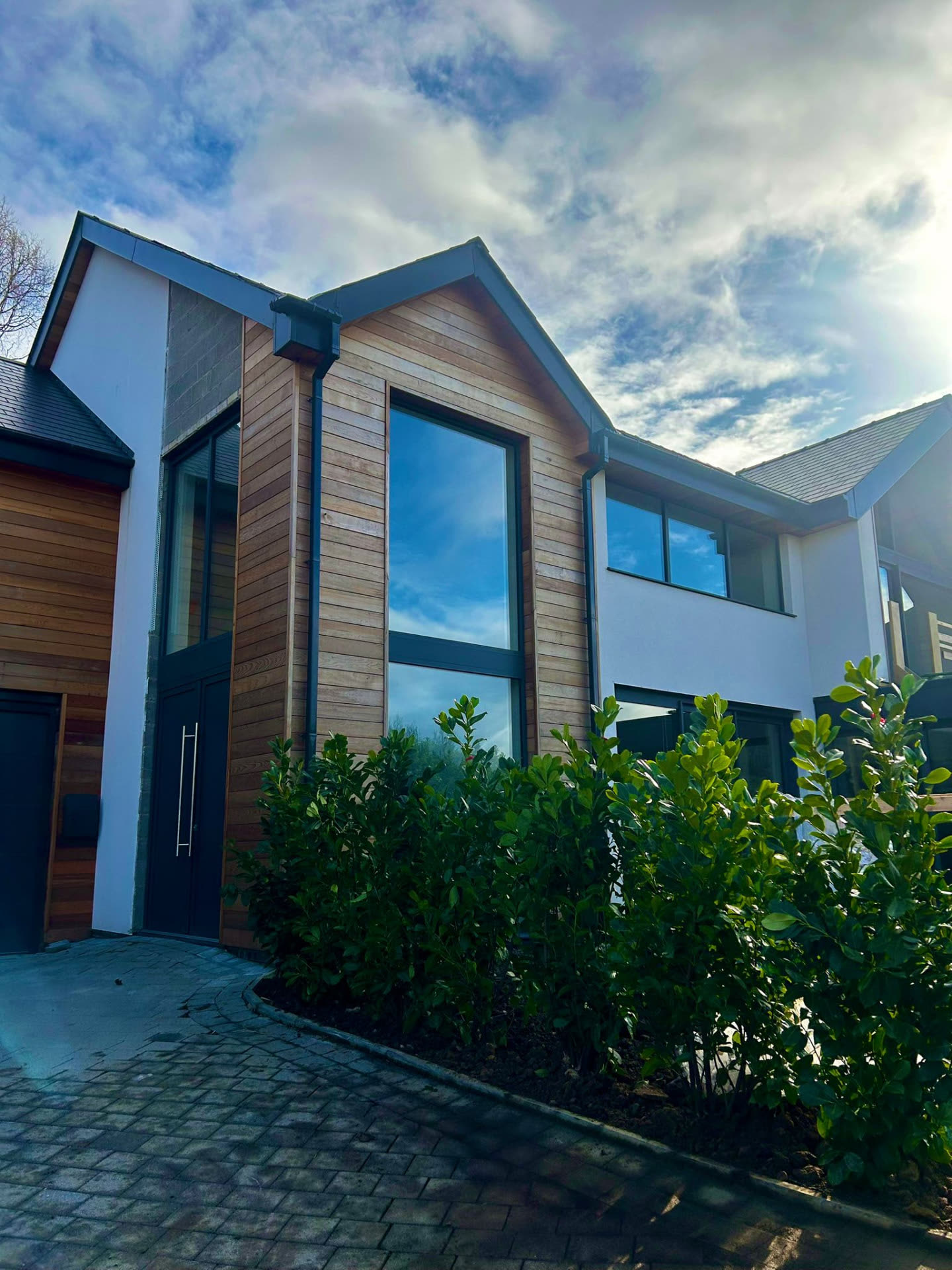 Modern timber-clad house entrance with tall glazing, dark front door and block paved driveway