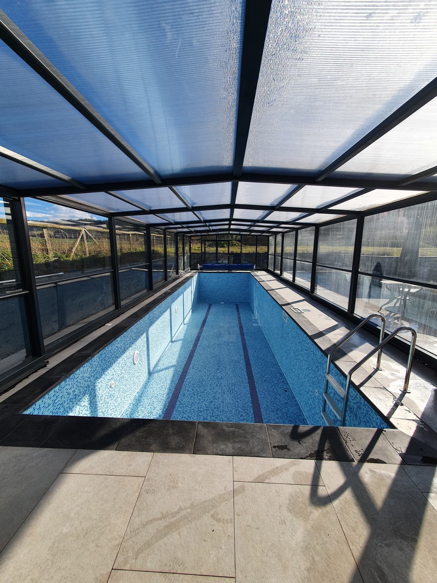 Long indoor swimming pool beneath a glass canopy roof with tiled poolside steps