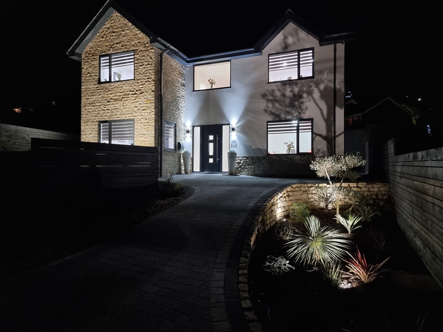 Two-storey stone-clad bespoke home lit at night with paved driveway and planted borders