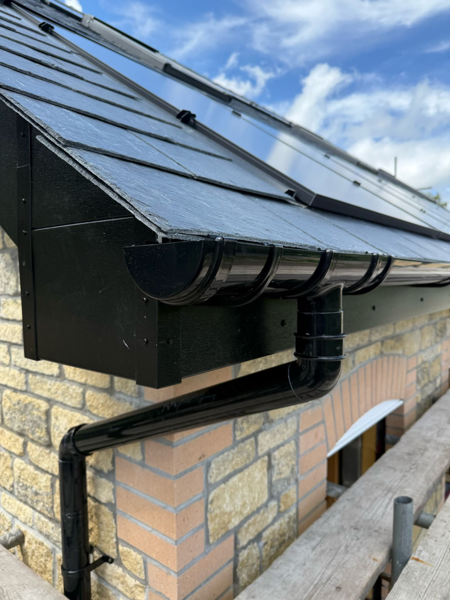 Detail of black guttering, fascia and downpipe against a slate roof and stone gable